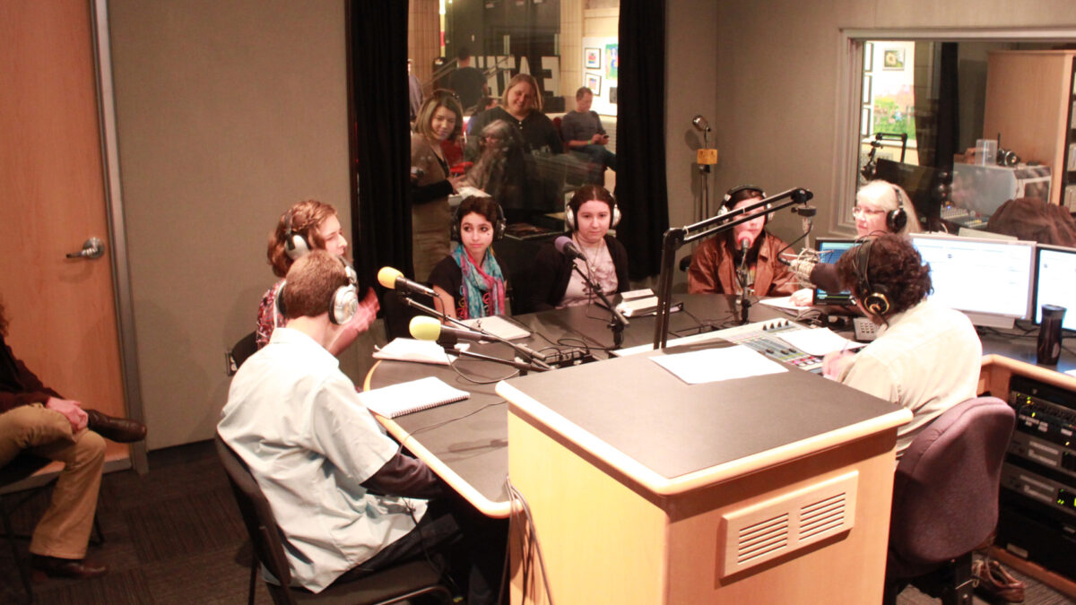 Five teens and an adult wearing headphones speaking into microphones being interviewed by an adult wearing headphones sitting behind a microphone and a mixer in a radio studio