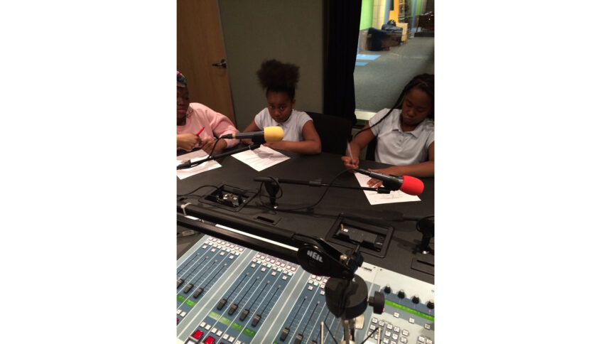 Three youth writing on pieces of paper sitting behind microphones in a radio studio