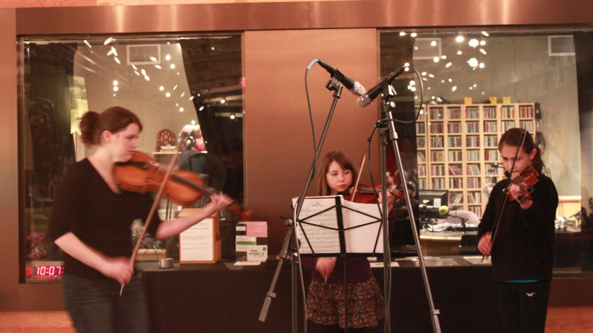 An adult and two youth playing violas looking at sheet music on a music stand in front of a radio studio