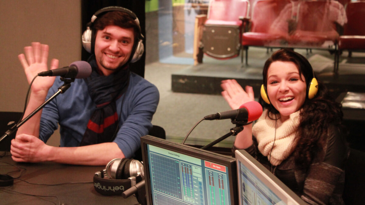 Two adults wearing headphones waving their hands behind microphones in a radio studio