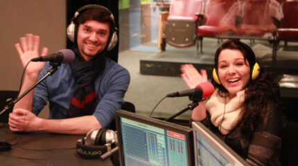 Two adults wearing headphones waving their hands behind microphones in a radio studio