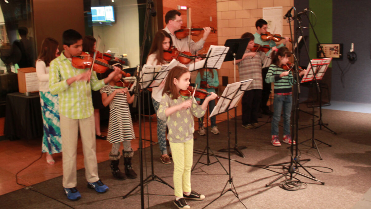Eight youth and an adult playing violins and looking at sheet music on music stands