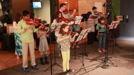 Eight youth and an adult playing violins and looking at sheet music on music stands