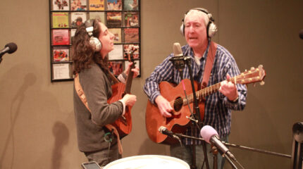 An adult wearing headphones playing ukulele and singing into a microphone and an adult wearing headphones playing acoustic guitar and singing into a microphone in a radio studio