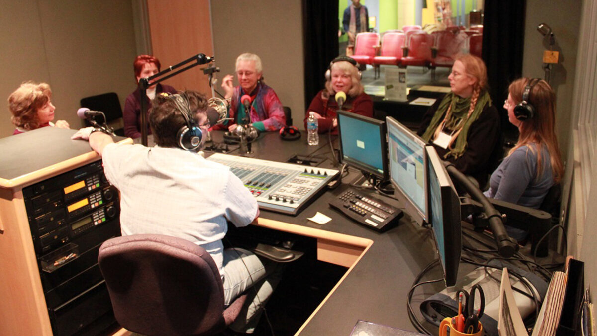 Six adults wearing headphones speaking into microphones being interviewed by an adult wearing headphones sitting behind a microphone and a mixer in a radio studio
