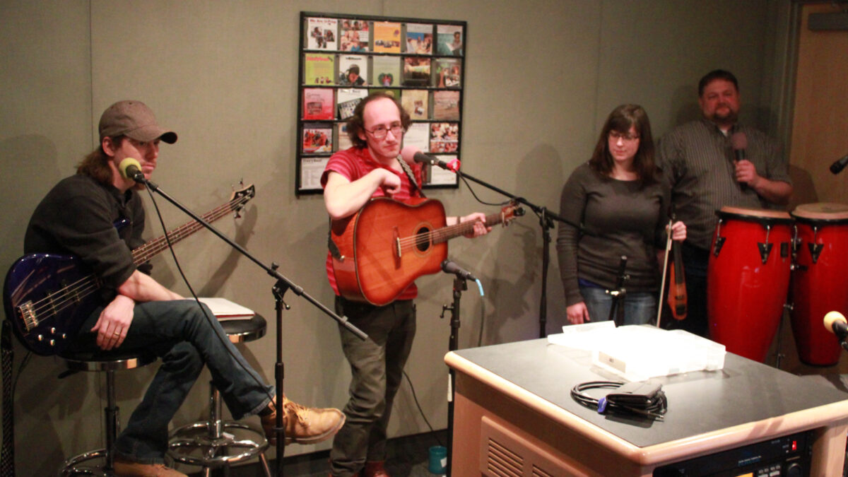 An adult holding an electric bass, an adult holding an acoustic guitar, an adult holding a violin, and an adult standing behind a pair of congas holding a microphone in a radio studio