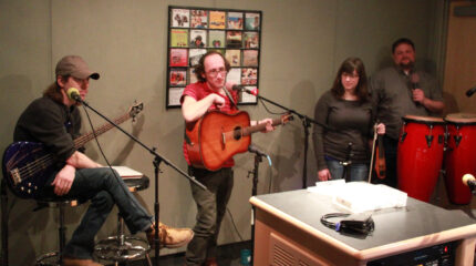 An adult holding an electric bass, an adult holding an acoustic guitar, an adult holding a violin, and an adult standing behind a pair of congas holding a microphone in a radio studio
