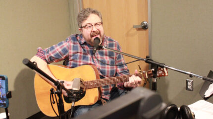 An adult playing an acoustic guitar and singing into a microphone in a radio studio