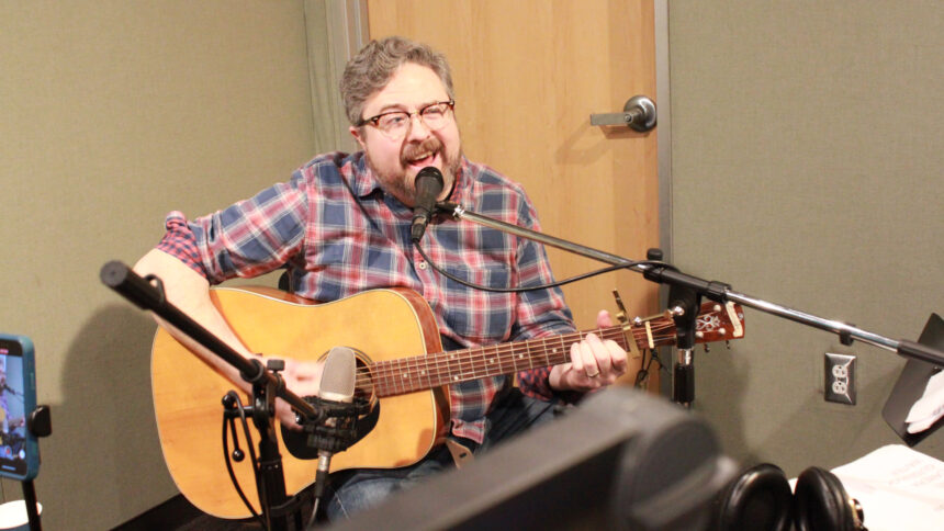 An adult playing an acoustic guitar and singing into a microphone in a radio studio