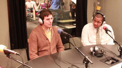 An adult and a teen wearing headphones speaking into microphones in a radio studio