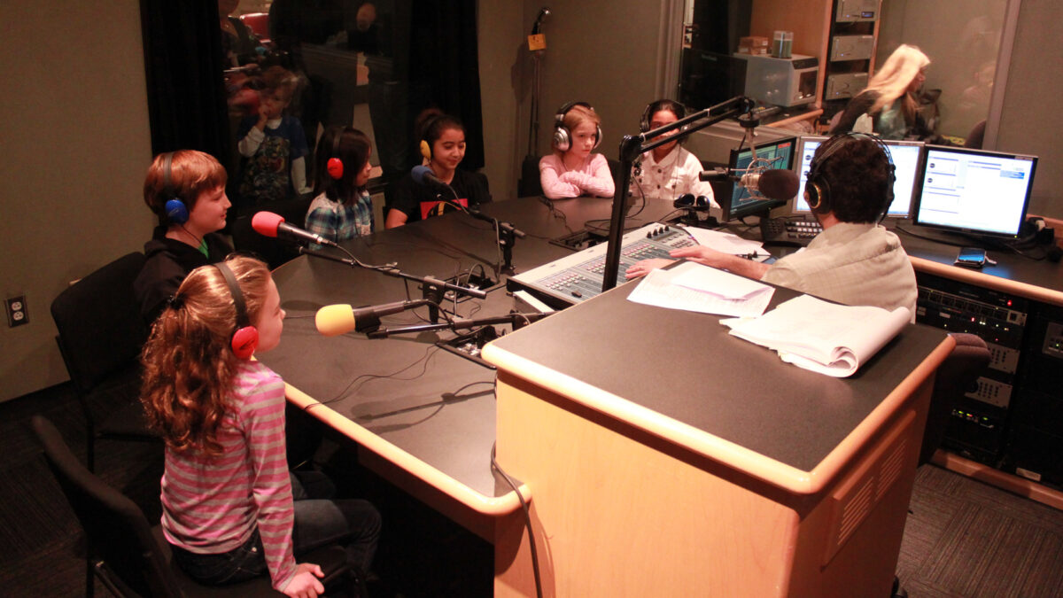 Six youth wearing headphones speaking into microphones being interviewed by an adult wearing headphones sitting behind a microphone and a mixer in a radio studio