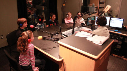 Six youth wearing headphones speaking into microphones being interviewed by an adult wearing headphones sitting behind a microphone and a mixer in a radio studio