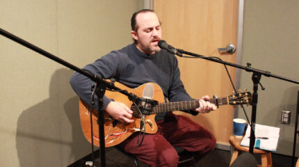 An adult playing acoustic guitar and singing into a microphone in a radio studio