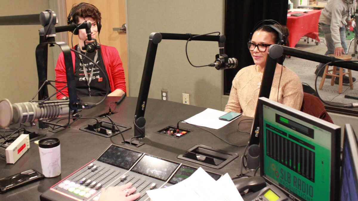 An adult and a teen wearing headphones speaking into microphones being interviewed in a radio studio