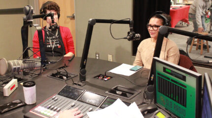 An adult and a teen wearing headphones speaking into microphones being interviewed in a radio studio