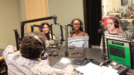 Three teens wearing headphones speaking into microphones being interviewed by an adult wearing headphones sitting behind a microphone and a mixer in a radio studio