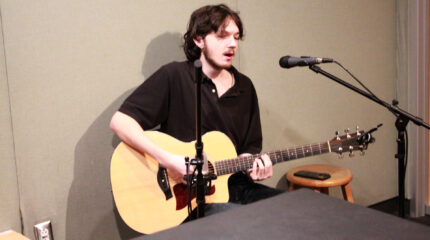 An adult playing acoustic guitar and singing into a microphone in a radio studio