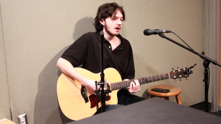 An adult playing acoustic guitar and singing into a microphone in a radio studio