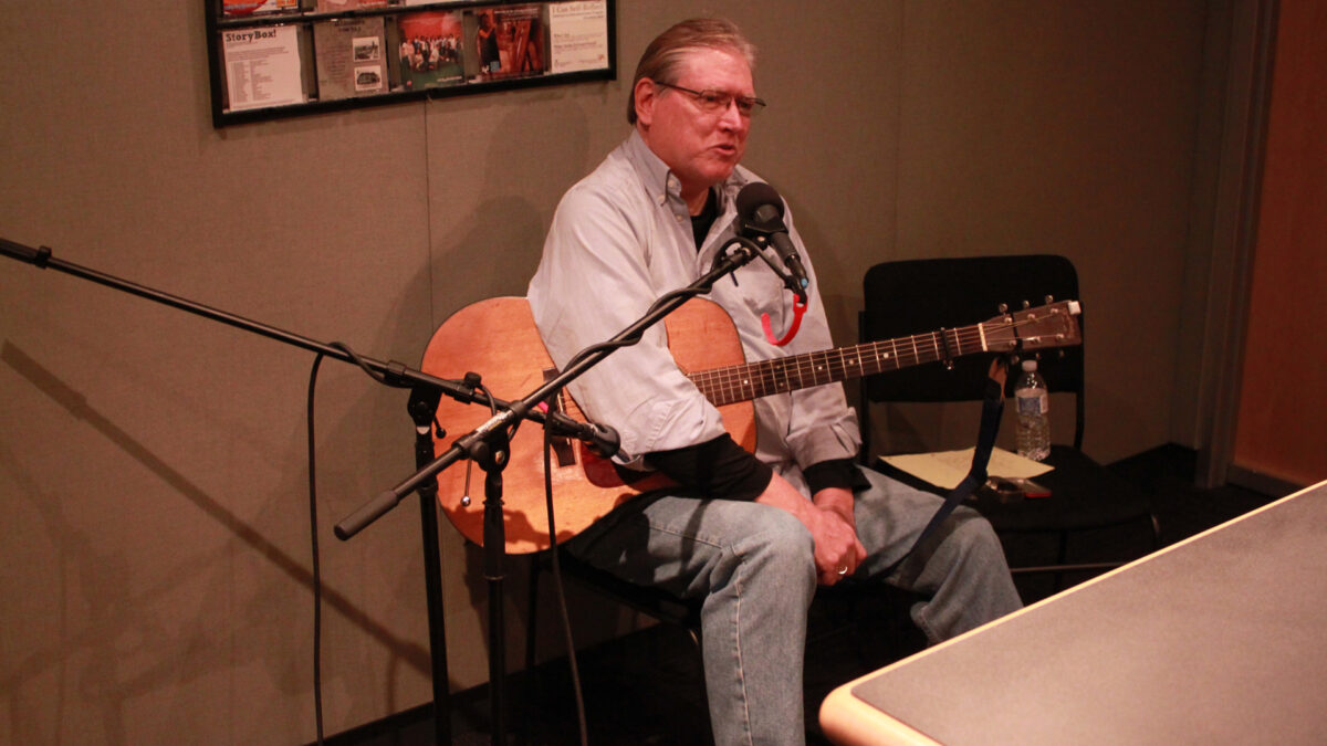 An adult sitting while holding an acoustic guitar and speaking into a microphone in a radio studio