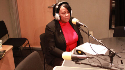 An adult wearing headphones sitting behind a microphone in a radio studio