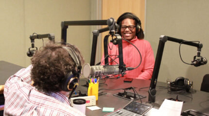 A teen wearing headphones smiling while sitting behind a microphone being interviewed by an adult wearing headphones sitting behind a microphone and a mixer in a radio studio