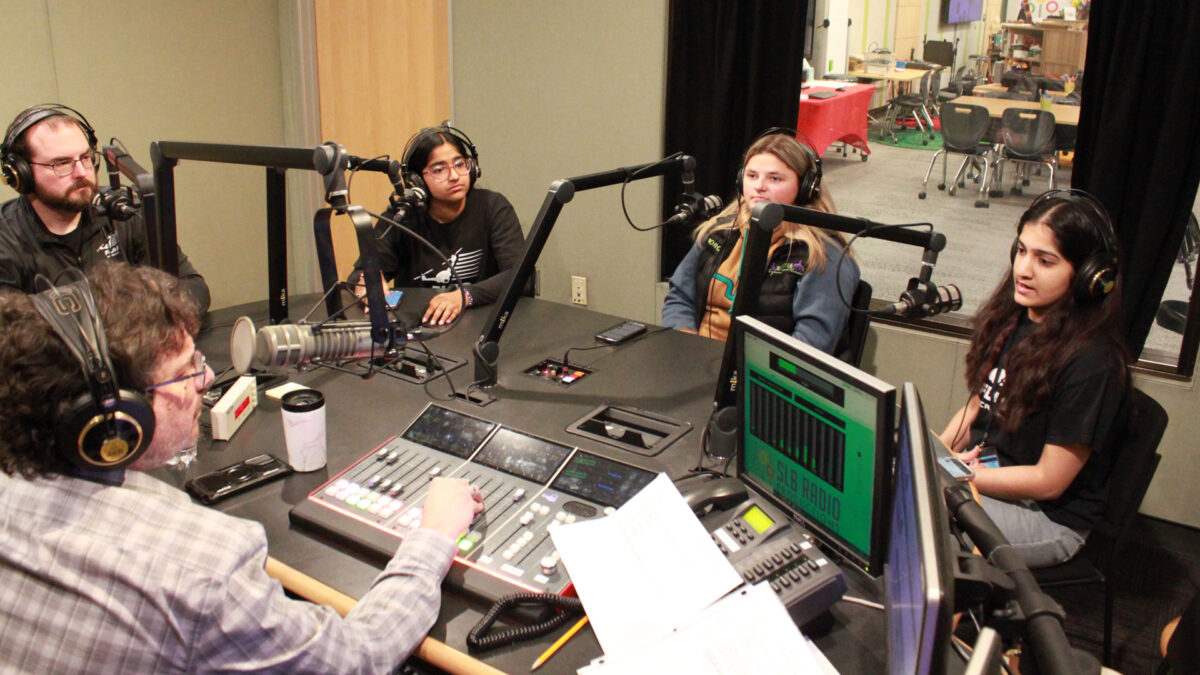 An adult and three teens wearing headphones speaking into microphones being interviewed by an adult wearing headphones sitting behind a microphone and a mixer in a radio studio