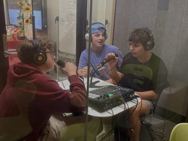 three teenagers in a glass booth with microphones, headphones, and an audio mixer