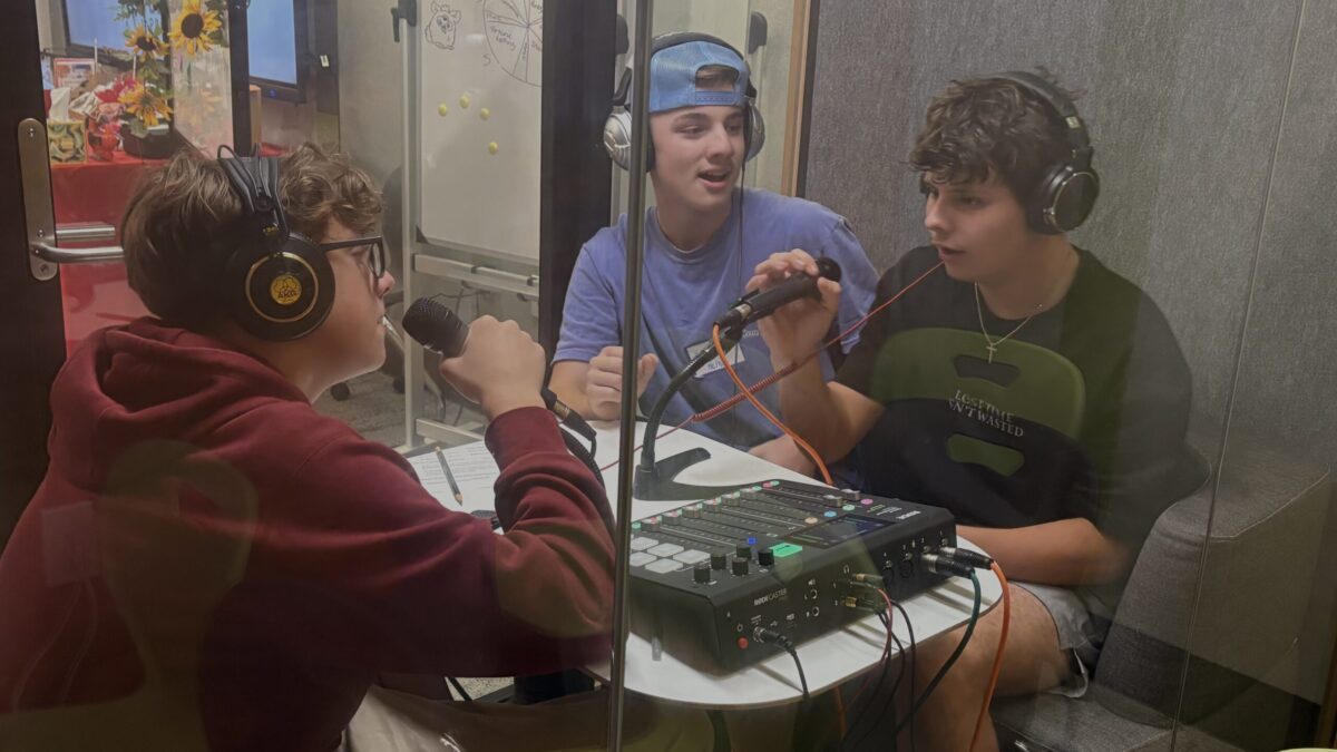 three teenagers in a glass booth with microphones, headphones, and an audio mixer