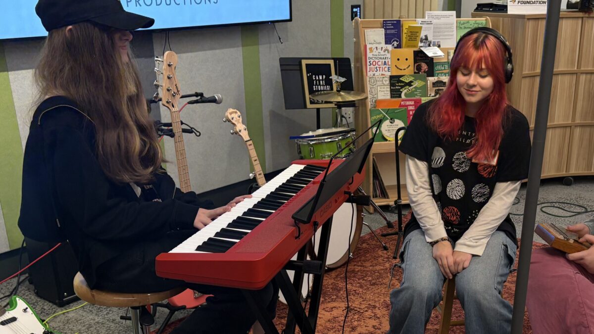 two teens sitting at a keyboard, one is playing the instrument, the other listens