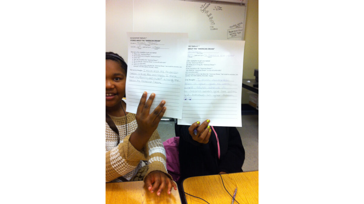 Two youth holding up pieces of paper to the camera while sitting at classroom desks