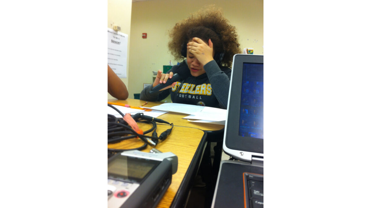 A youth wearing headphones speaking into a microphone while sitting at a classroom desk