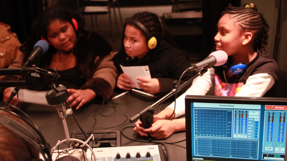 Three youth wearing headphones speaking into microphones and holding pieces of paper in a radio studio