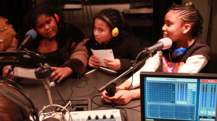 Three youth wearing headphones speaking into microphones and holding pieces of paper in a radio studio