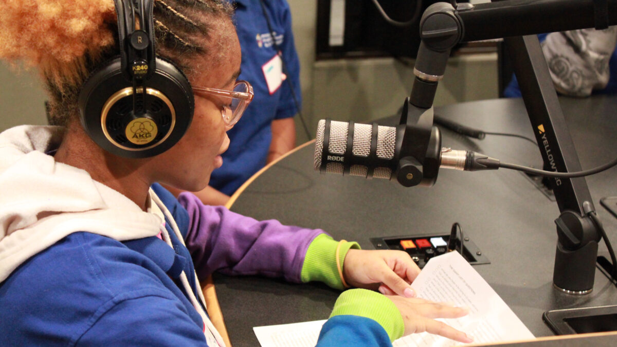 A teen wearing headphones sitting behind a microphone and looking down at a piece of paper in a radio studio