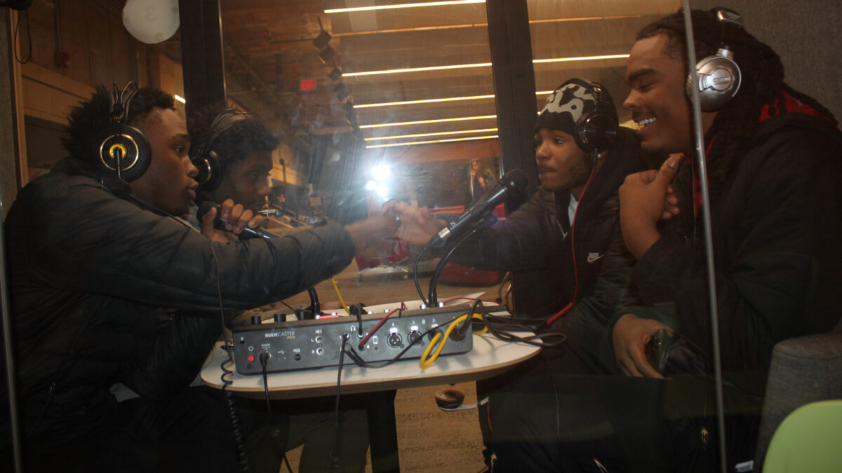 Four teens wearing headphones speaking into microphones in a recording booth