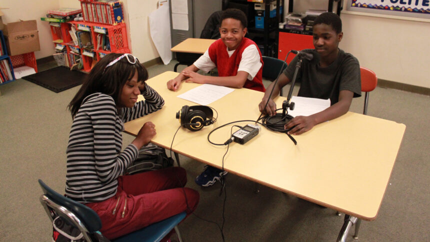 Three youth sitting around a microphone and a portable recorder on a classroom desk