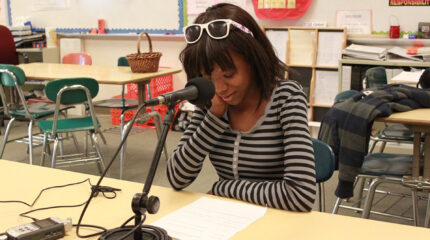A teen sitting behind a microphone and a portable recorder looking at a piece of paper on a classroom table