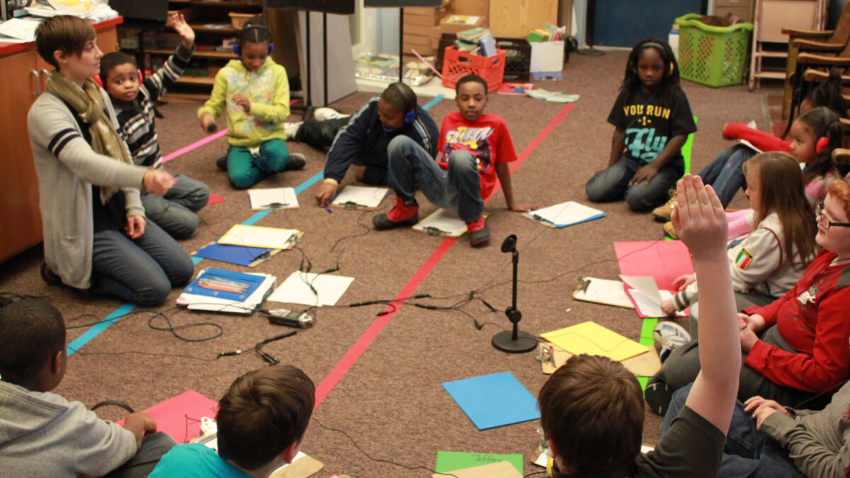 Twelve youth, five wearing headphones, and an adult all sitting in a circle on the floor around a microphone and a portable recorder