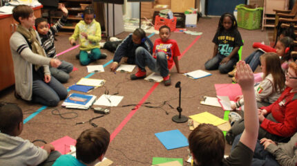 Twelve youth, five wearing headphones, and an adult all sitting in a circle on the floor around a microphone and a portable recorder