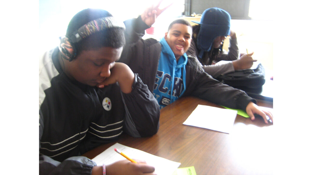 Two youth, one wearing headphones, writing on pieces of paper and a youth smiling at the camera and flashing a peace sign