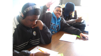 Two youth, one wearing headphones, writing on pieces of paper and a youth smiling at the camera and flashing a peace sign