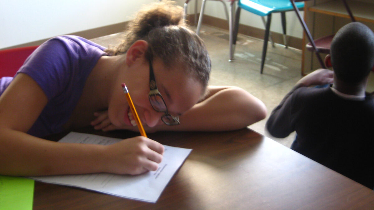 A youth smiling while writing on a piece of paper on a classroom desk and a youth sitting on the floor in a classroom