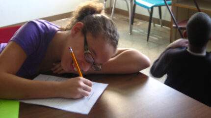 A youth smiling while writing on a piece of paper on a classroom desk and a youth sitting on the floor in a classroom