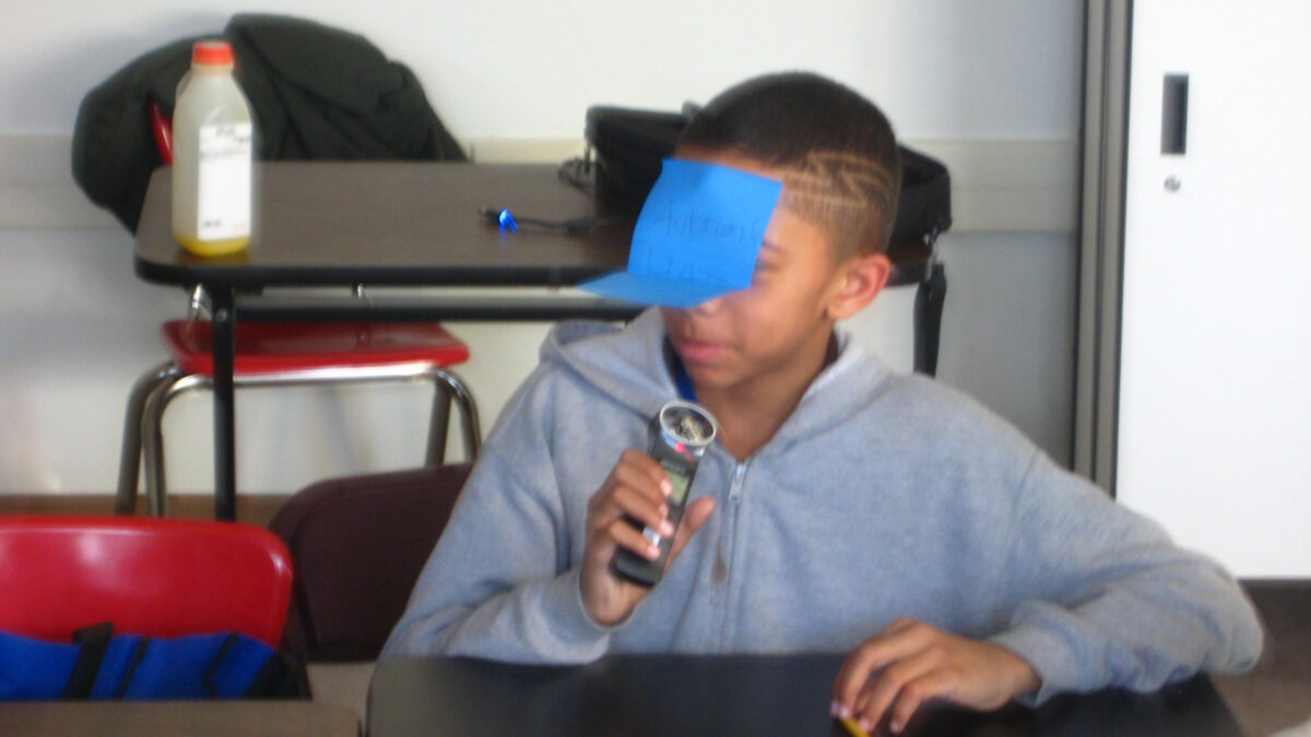 A youth with a sticky note on their face speaking into a portable recorder while sitting at a classroom desk