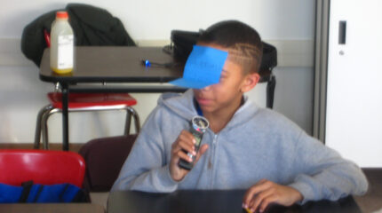 A youth with a sticky note on their face speaking into a portable recorder while sitting at a classroom desk