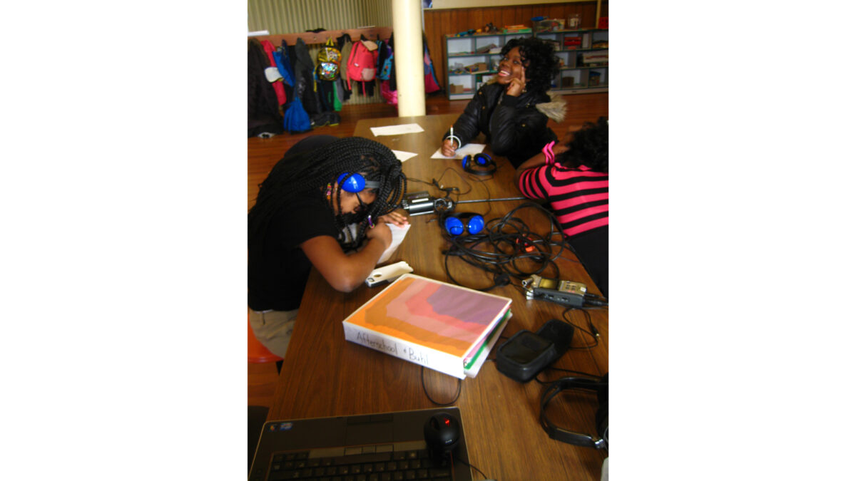 Three youth, one wearing headphones, writing on pieces of paper sitting at wooden tables