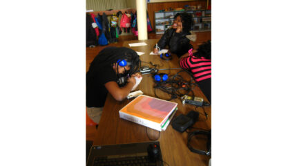 Three youth, one wearing headphones, writing on pieces of paper sitting at wooden tables