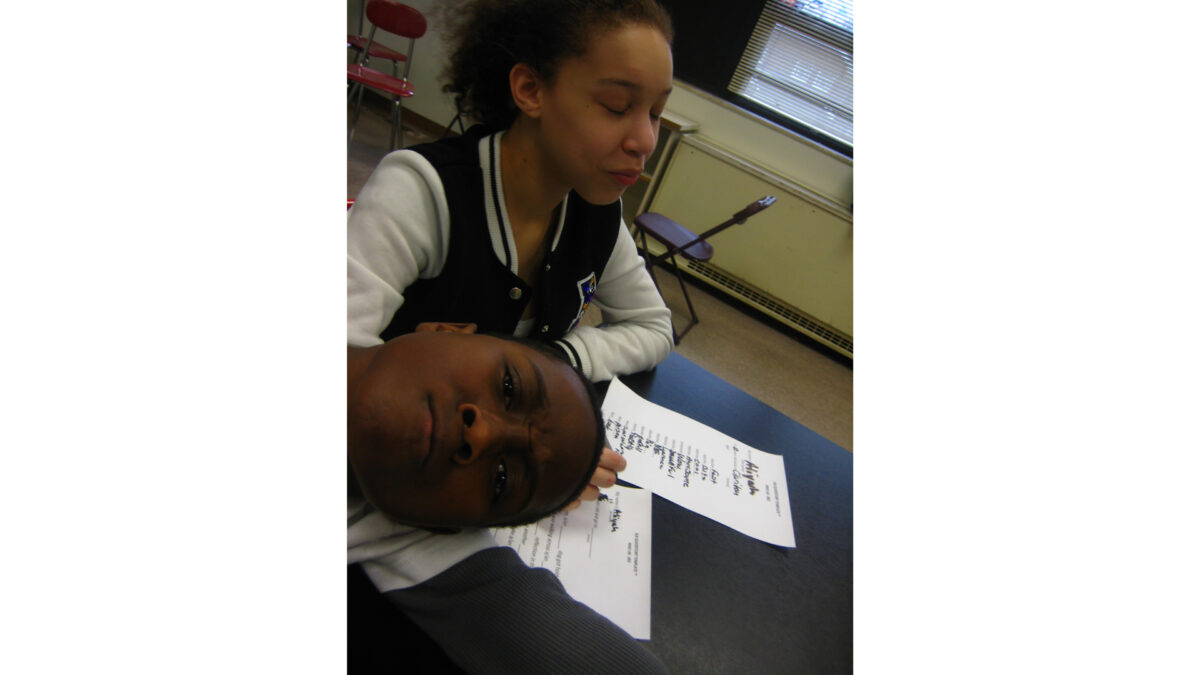 Two youth making faces at the camera while sitting behind pieces of paper on a classroom desk
