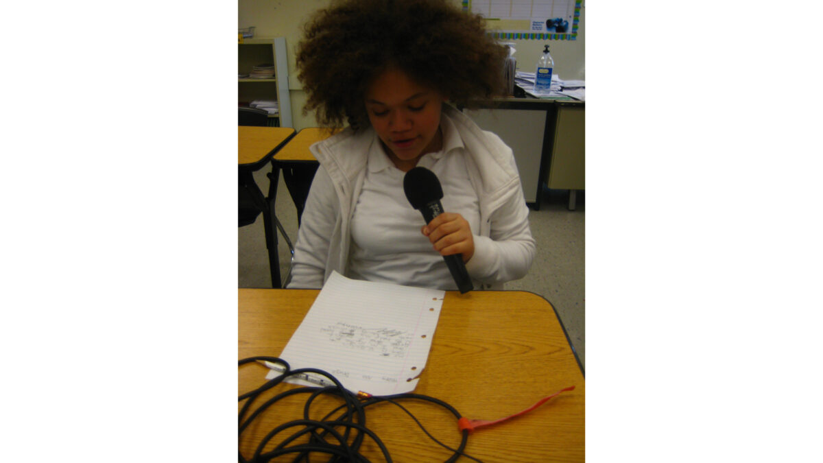 A youth holding and speaking into a microphone while looking down at a piece of paper on a classroom desk