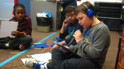 Three youth wearing headphones sitting on the floor, one holding a piece of paper, and one holding and looking down at a clipboard and speaking into a microphone